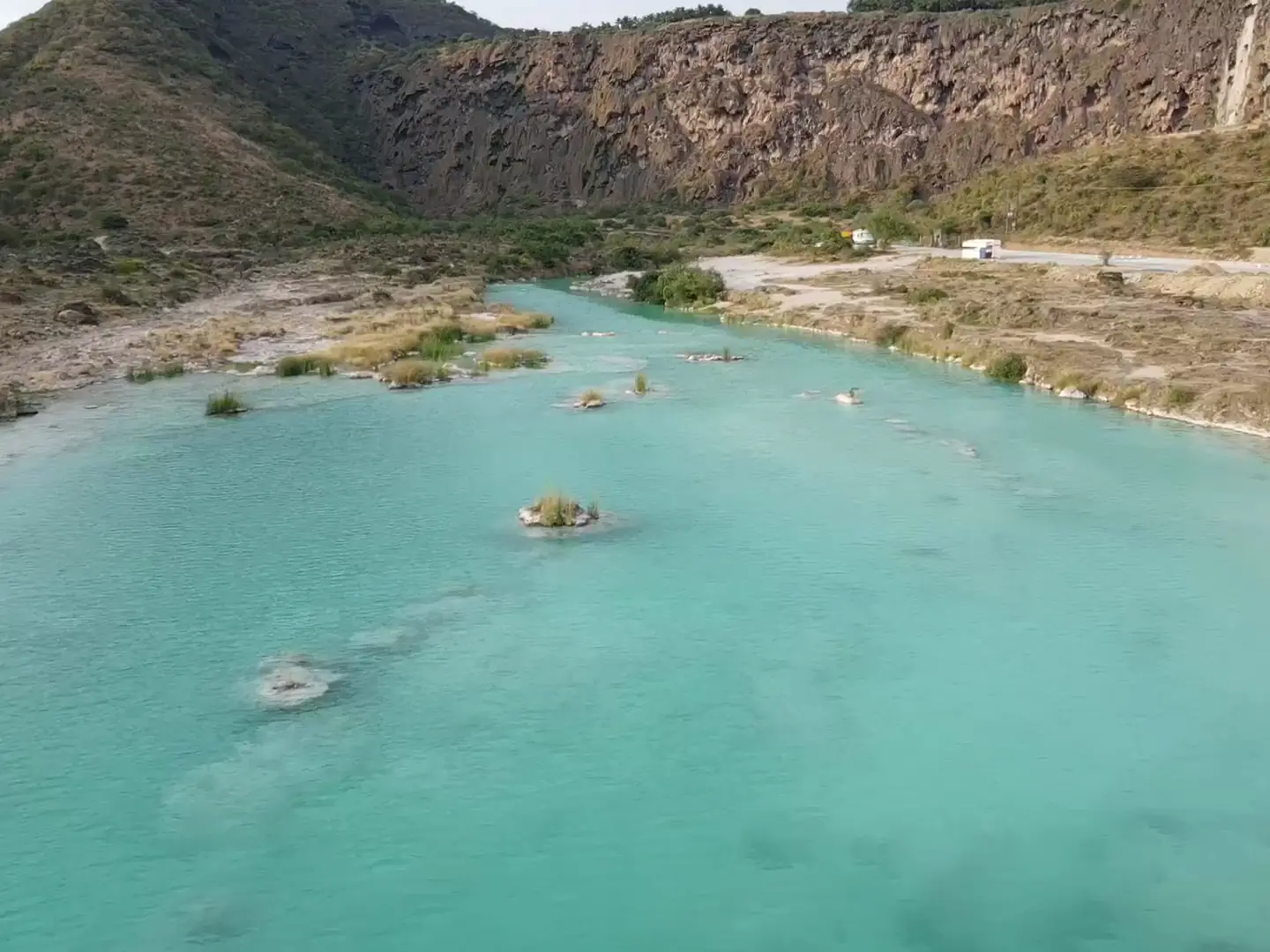 Film flying low over the turquoise lake of Wadi Darbat, a waterfall spilling over travertine rock into a pool, and khareef mist over green Dhofar hills